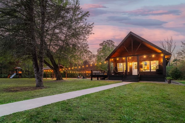 Illuminated log cabin at dusk with firepit seating area, string lights, and surrounding trees