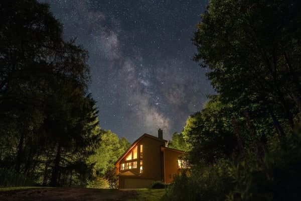 Rustic log cabin with covered porch surrounded by tall evergreen forest and gravel driveway
