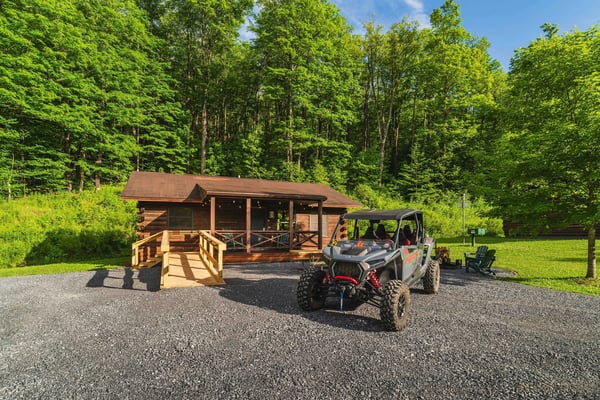 Wooden cabin with covered porch and ramp surrounded by dense forest, with off-road vehicle parked in gravel driveway