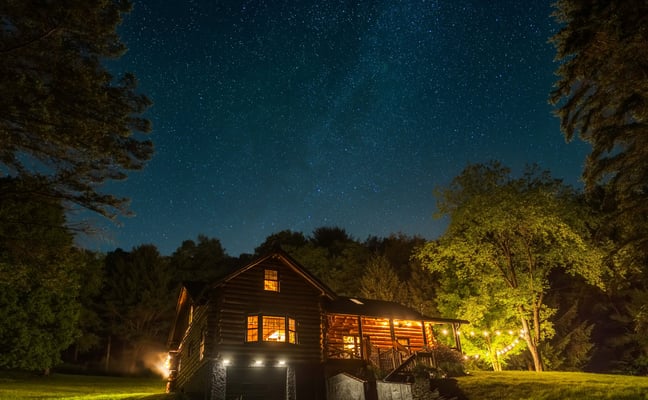 Illuminated log cabin in forest clearing at night with starry sky and tall trees surrounding property